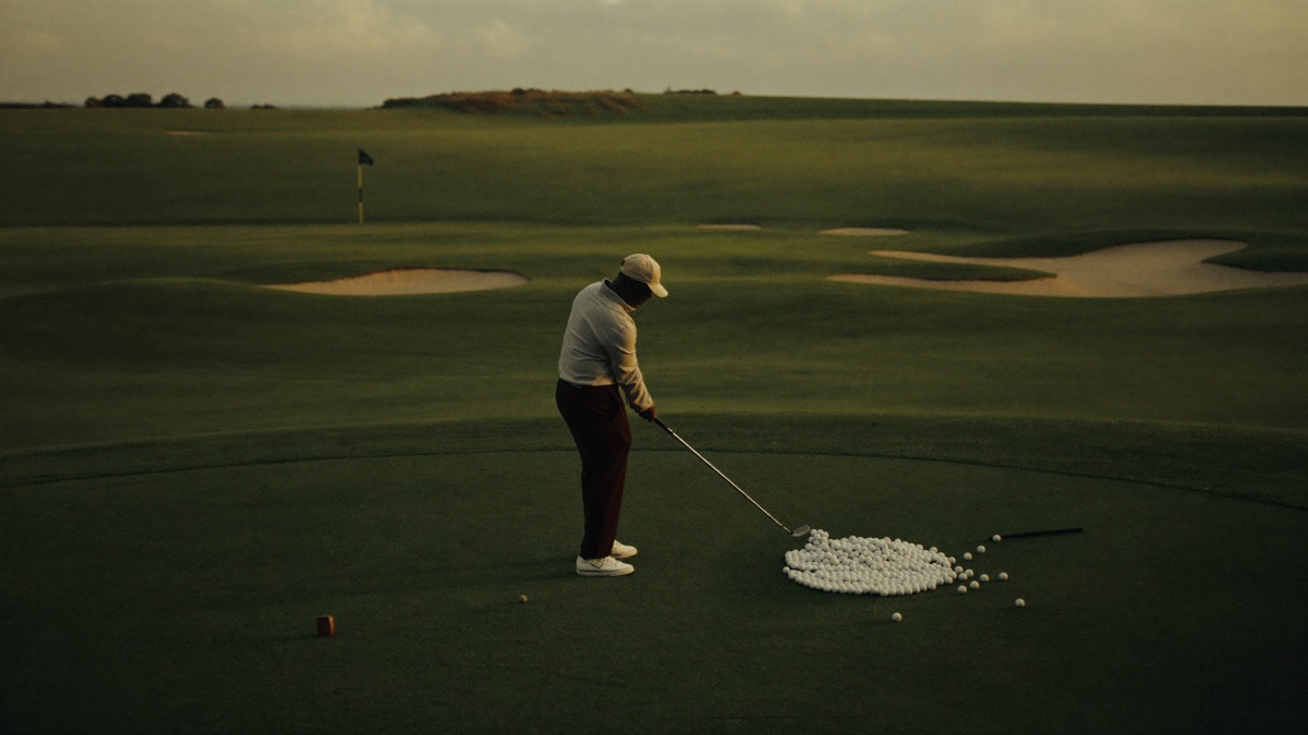 A lone golfer at dusk standing over a large scattered pile of practice balls on an empty practice ground, long shadows across the turf