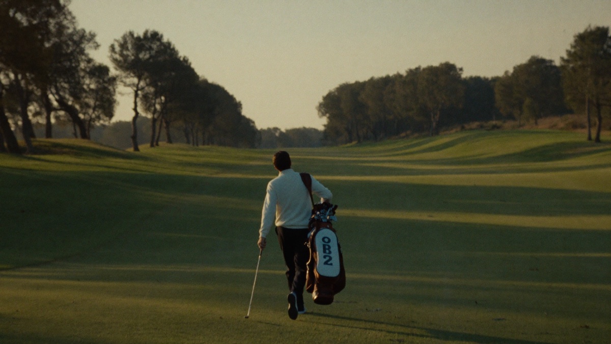 A golfer seen from behind carrying a stand bag up the middle of a wide striped fairway at golden hour, long tree shadows across the grass and a distant green ahead