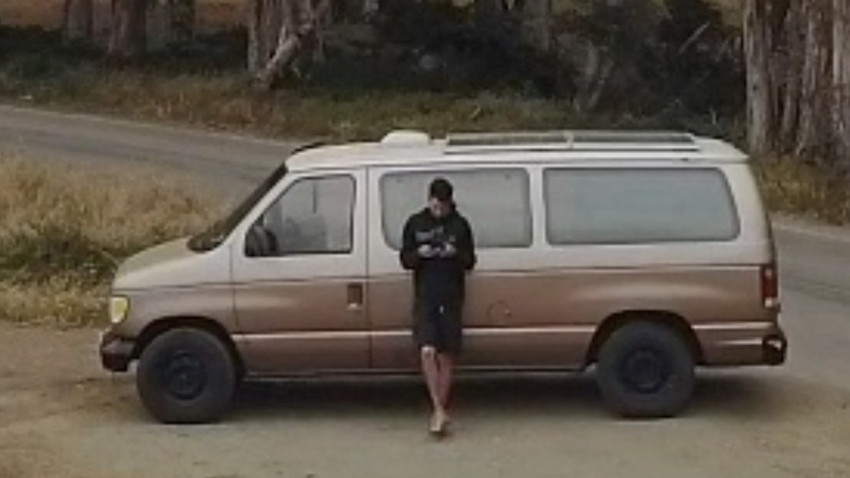 Cameron Tennant leaning against the side of the tan Ford van he lived in, parked on a dirt road lined with eucalyptus trees