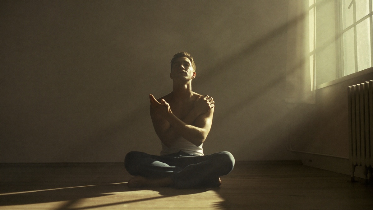 A calm man seated cross-legged on a wooden floor in golden dawn light, eyes closed and face lifted toward the window, arms crossed over his chest mid slow breath