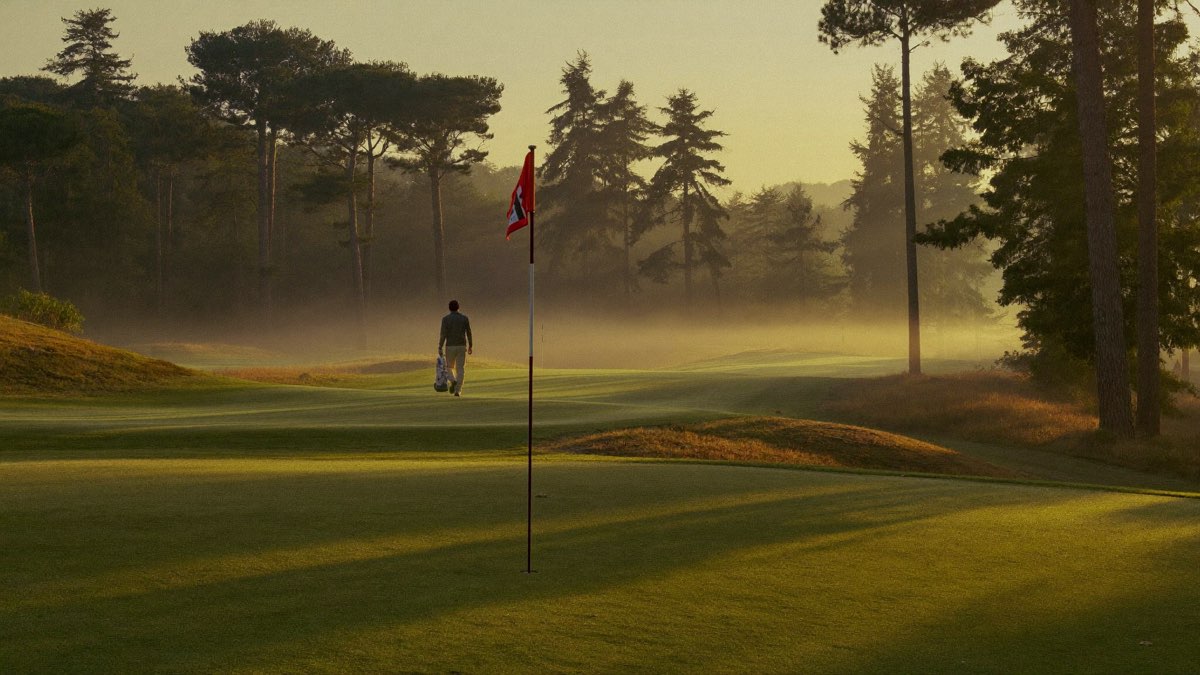 A lone golfer carrying a light bag walks toward a close red flagstick through sunrise mist on a quiet 9-hole par 3 course