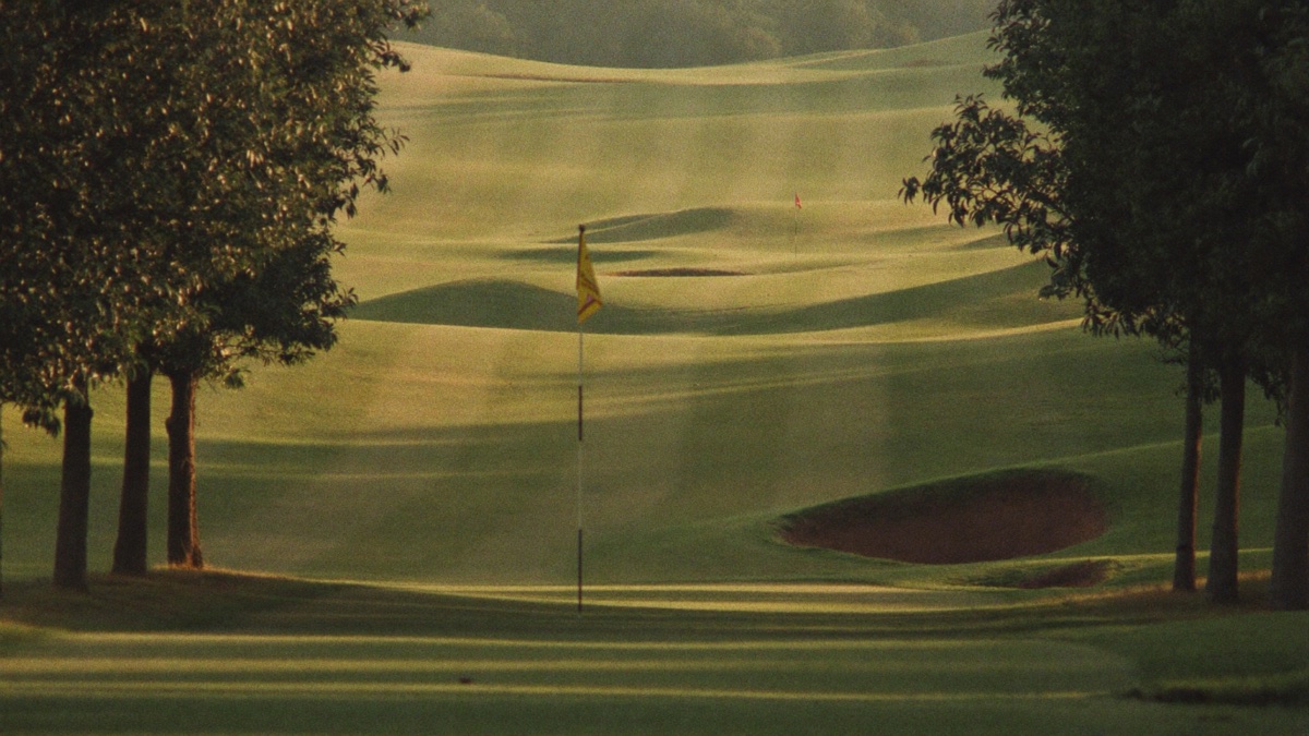 A quiet tree-lined golf hole in soft evening light, a yellow flag standing on the green in the foreground and mown fairway rolling away past a deep bunker toward a second flag in the distance