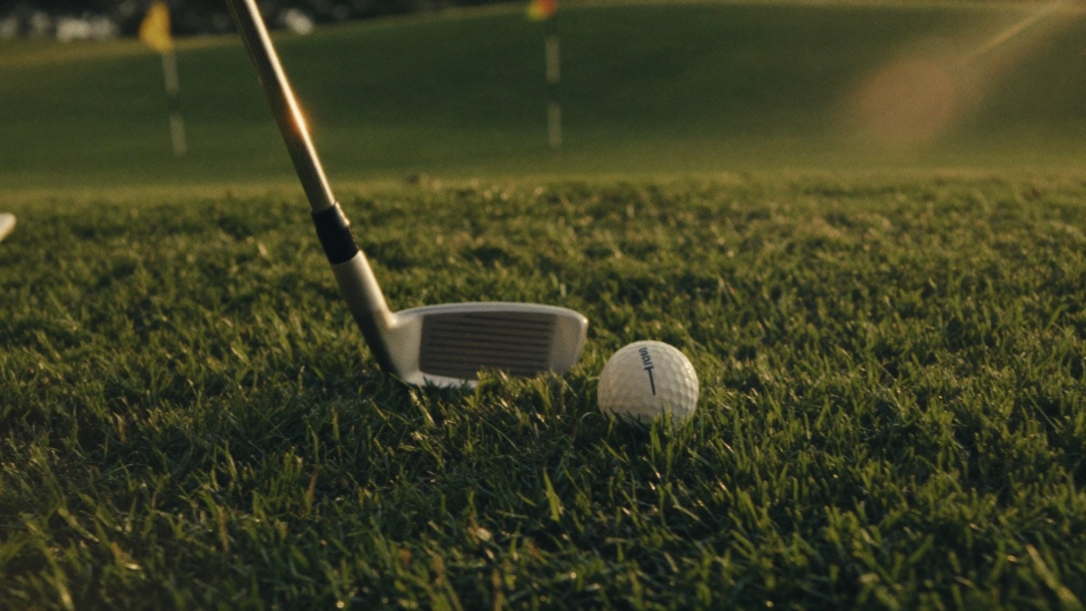 Close-up of a golf ball sitting in the grass with the head of an iron grounded just behind it, low golden light raking across the blades and a distant green and flagsticks blurred beyond