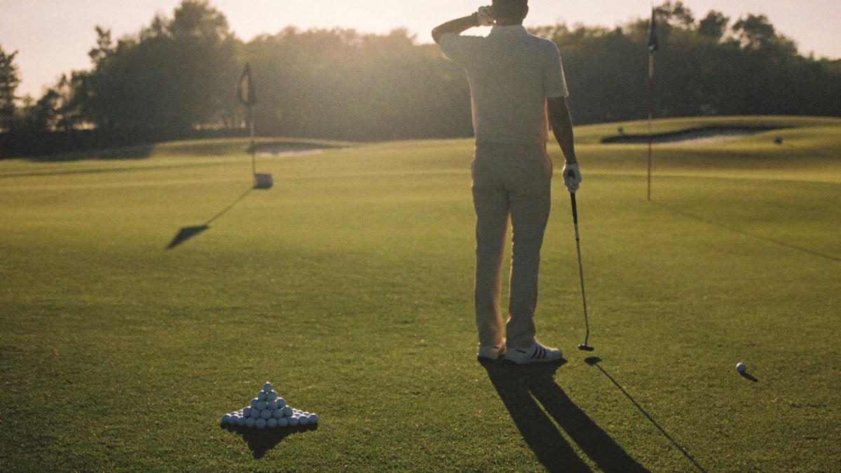 A lone golfer at golden hour on an empty practice ground, a neat pyramid of balls on the turf beside him and an iron grounded at his side, his shadow stretching long across the grass