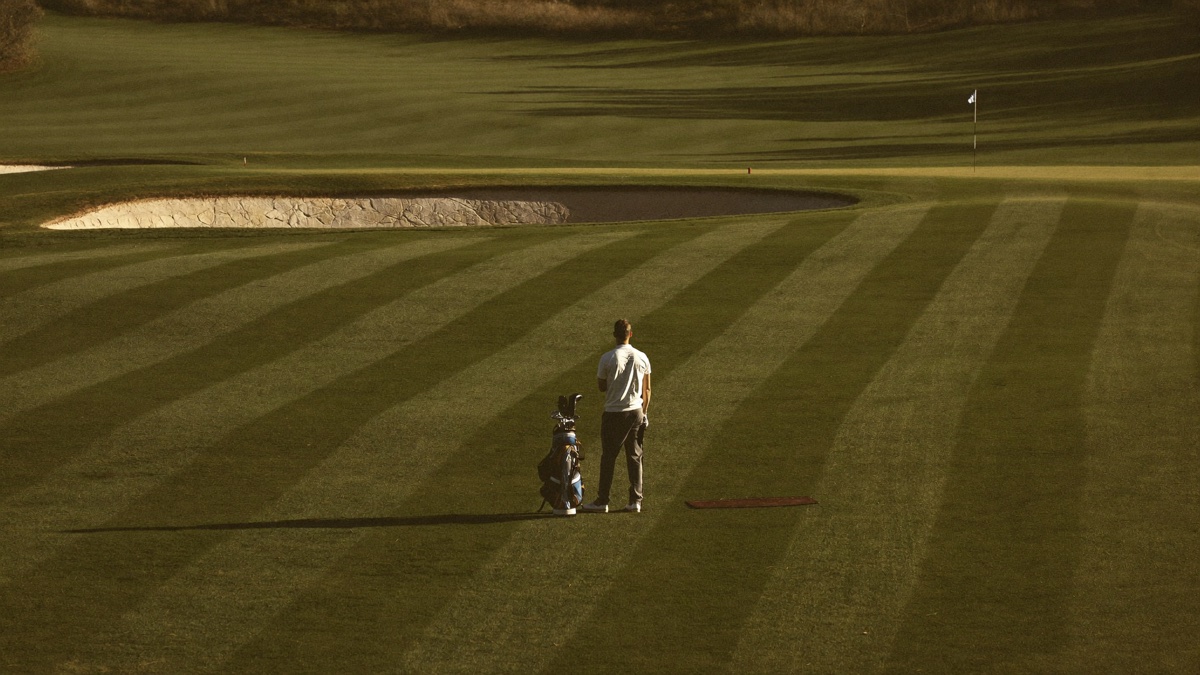 A golfer standing alone in the middle of a wide striped fairway at golden hour, his bag on the grass beside him, looking up the hole toward a distant green and a cluster of bunkers