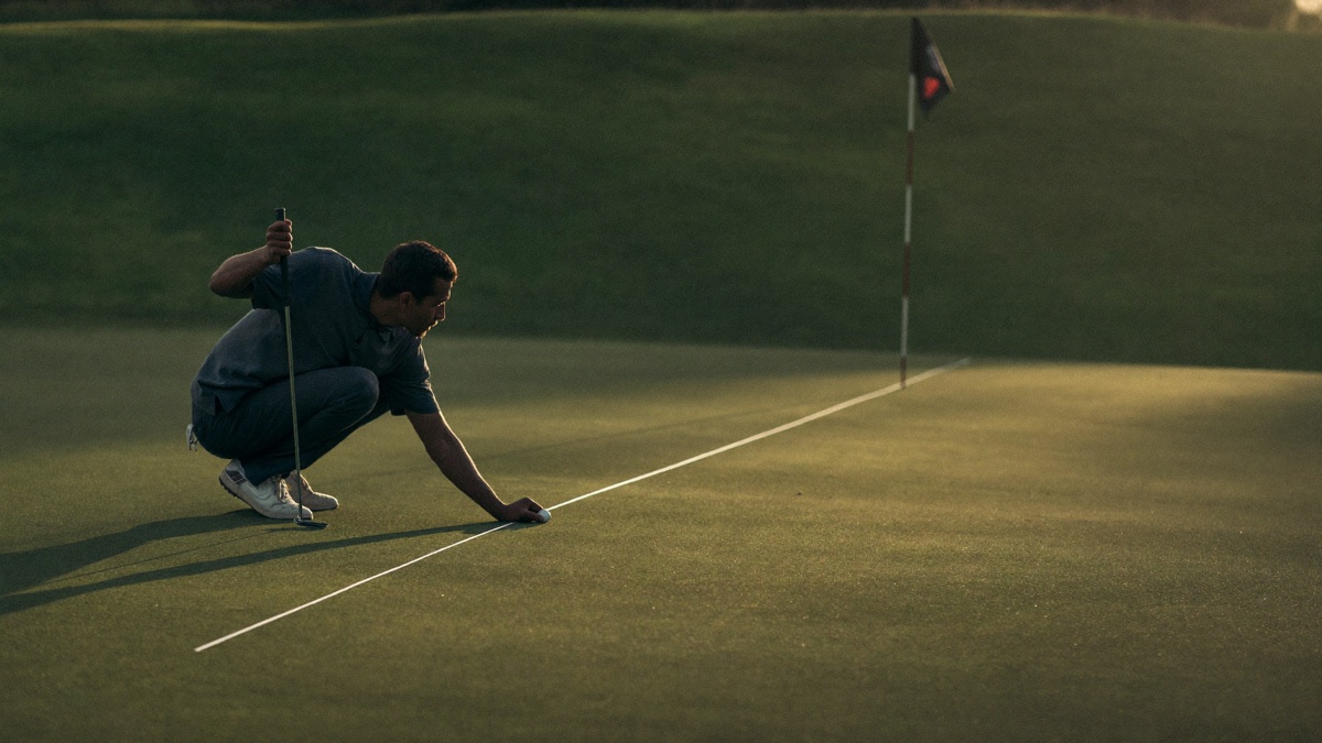 Low wide-angle view of a golfer crouched behind his ball reading the line of a long lag putt on a large green at golden hour, putter in hand, flagstick roughly forty feet away