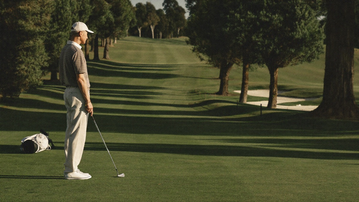A golfer standing alone in a tree-lined fairway at golden hour, an iron resting head-down on the grass and his bag on the ground behind him, looking down the hole toward a distant bunker