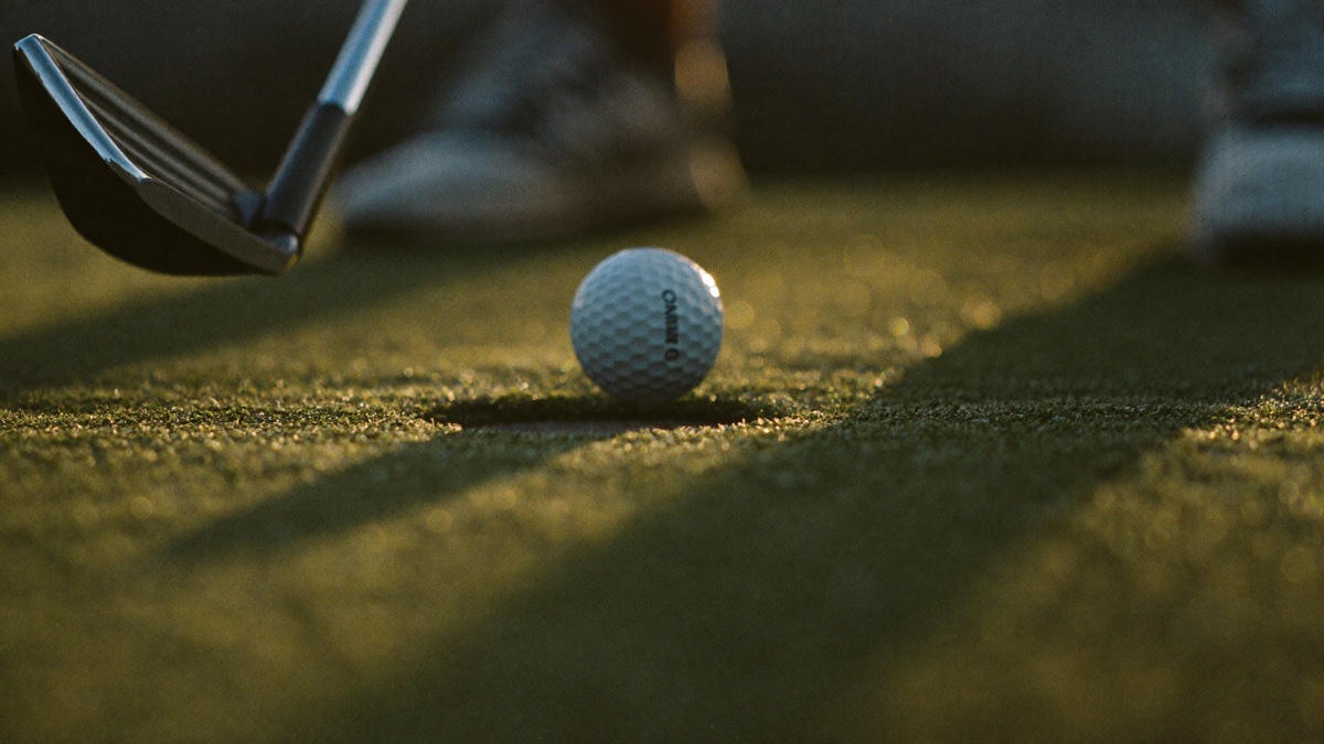 Low-angle close-up of a golf ball sitting on a green in low golden light, the head of a putter and the golfer's shoes softly blurred behind it