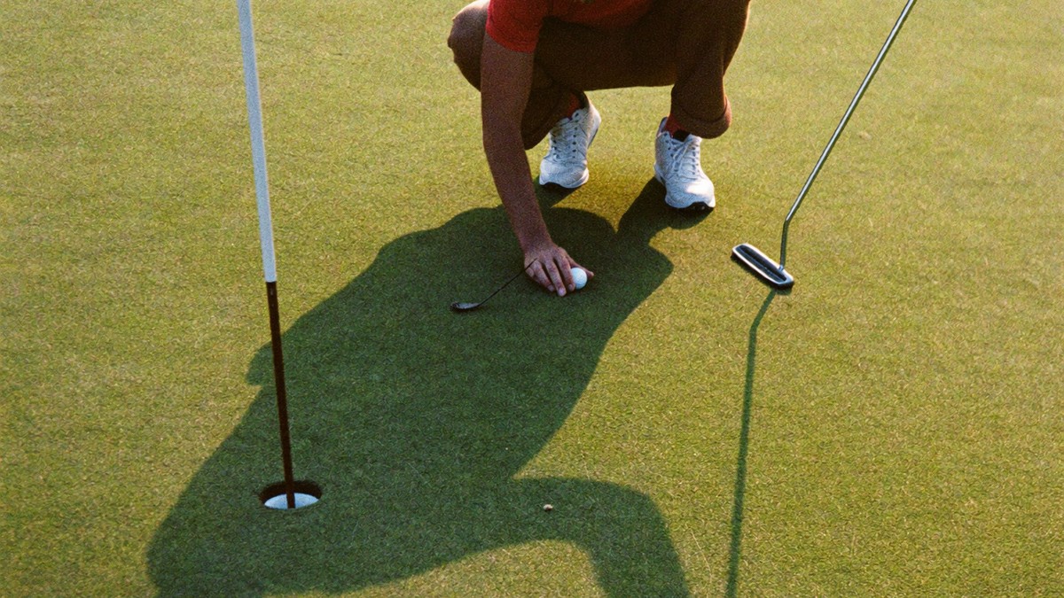 A golfer crouches to mark his ball on the green at golden hour, the ball resting just a couple feet from the hole with the flagstick and his putter beside it