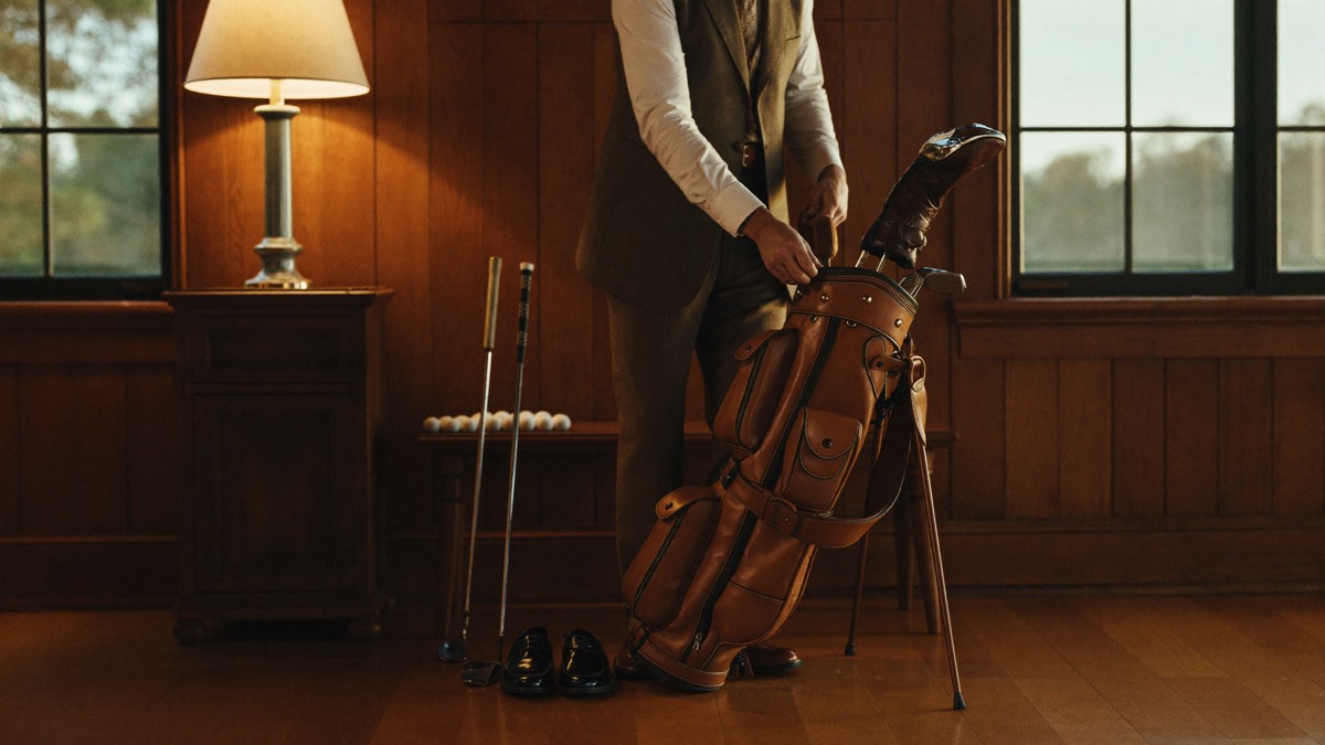 A golfer in classic attire packing a vintage tan leather golf bag in a warm lamplit wood-paneled room the night before a round, golf balls lined up on a bench beside polished leather shoes
