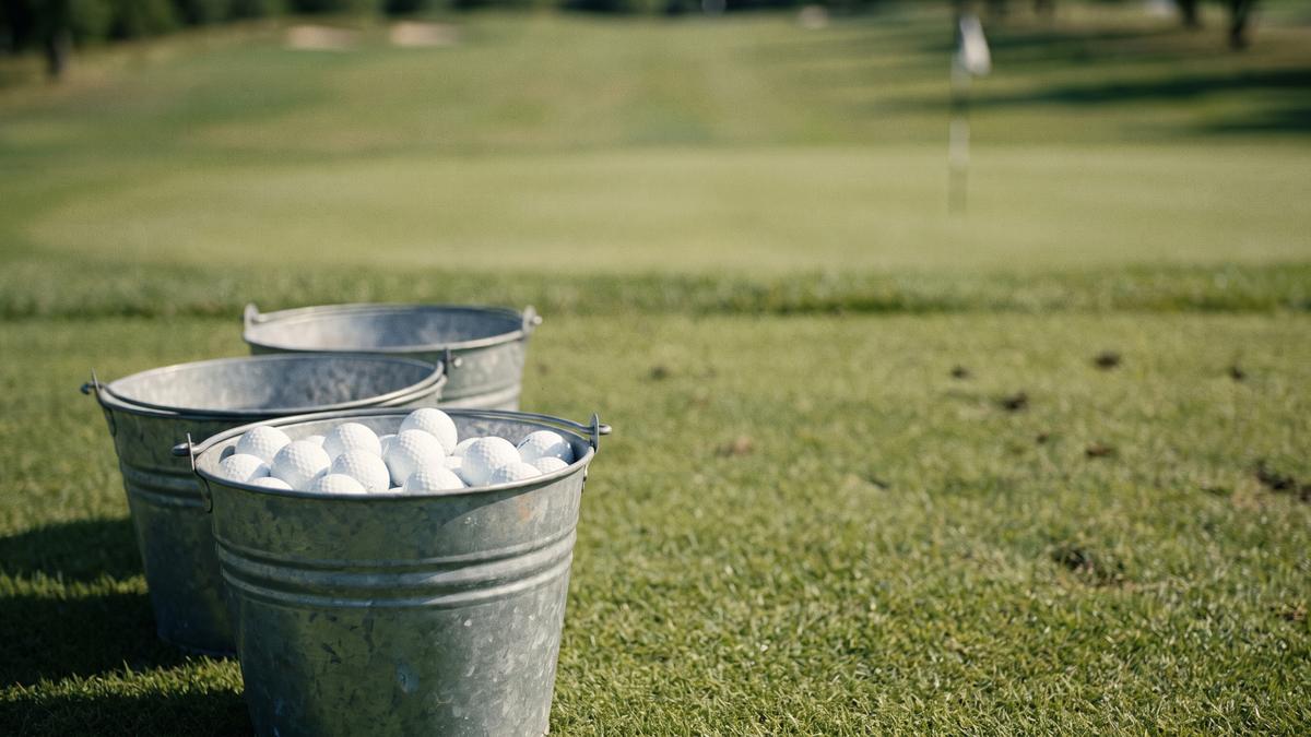 Three metal range buckets on a practice tee in evening sun, only the nearest one filled with golf balls, with a fairway and a flag on a distant green behind them