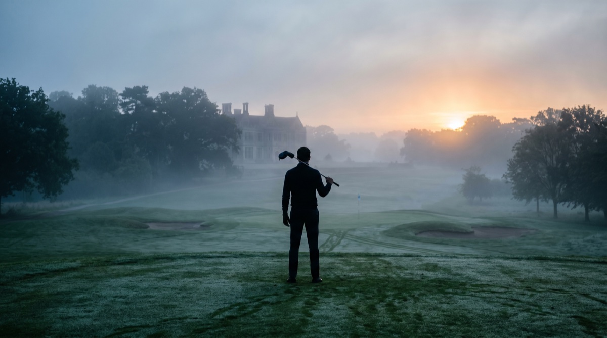 A golfer standing alone in morning mist at dawn, driver over his shoulder, looking down the first fairway toward an old clubhouse at sunrise