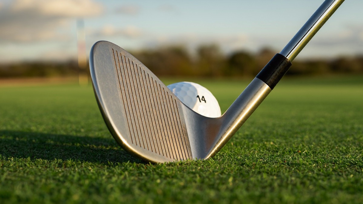 A single lofted sand wedge at address on mown fairway turf behind a white golf ball at golden hour, with an out-of-focus flagstick beyond