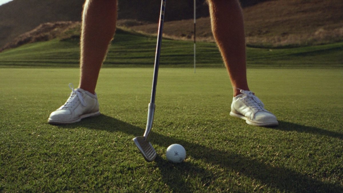 Low-angle close-up of a golfer's shoes and legs settled in a balanced stance on mown fairway grass beside a golf ball, warm light raking across the turf with a distant flag out of focus behind