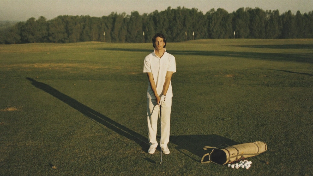 A lone golfer standing completely still on a wide open practice field at golden hour, an iron grounded in front of him and a tipped-over canvas shag bag of balls at his feet, small target flags along the distant tree line