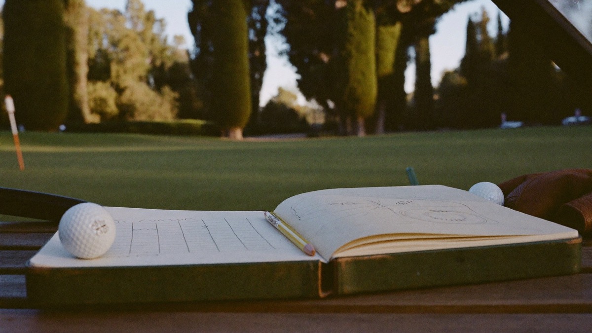 An open golf journal with a pencil, a hand-drawn grid and a sketched green on its pages, resting on a wooden bench beside golf balls and a leather glove, a tree-lined fairway in warm evening light behind